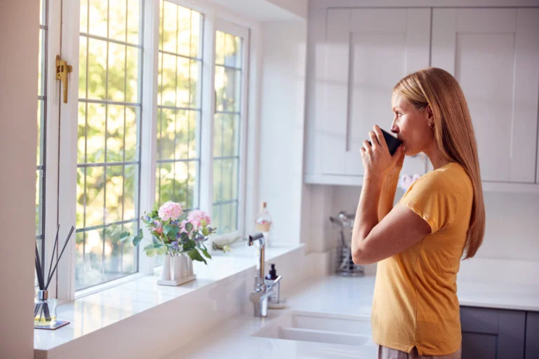 Mature Woman At Home With Hot Drink Standing By Kitchen Window